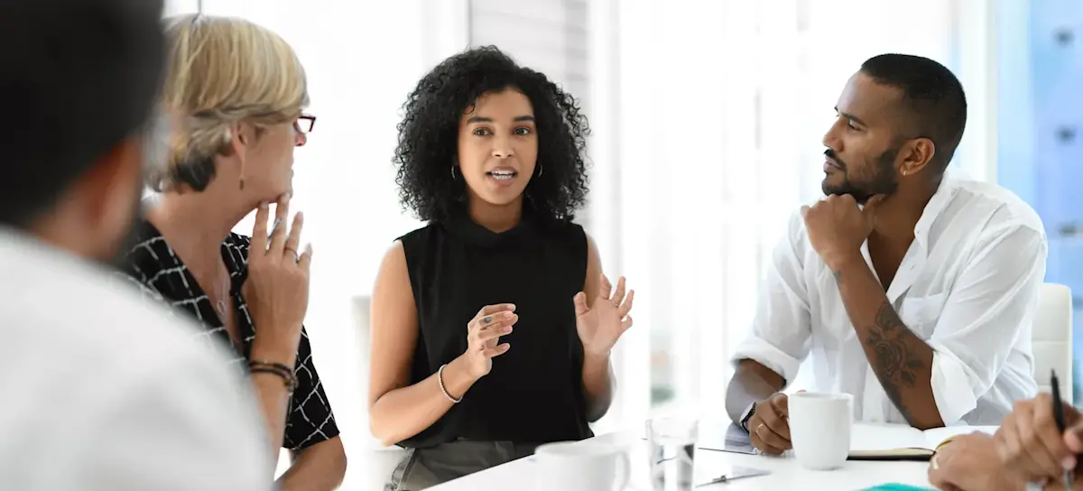 [Featured image] A group of colleagues sits around, actively listening to a person in the middle of the conference table speak.