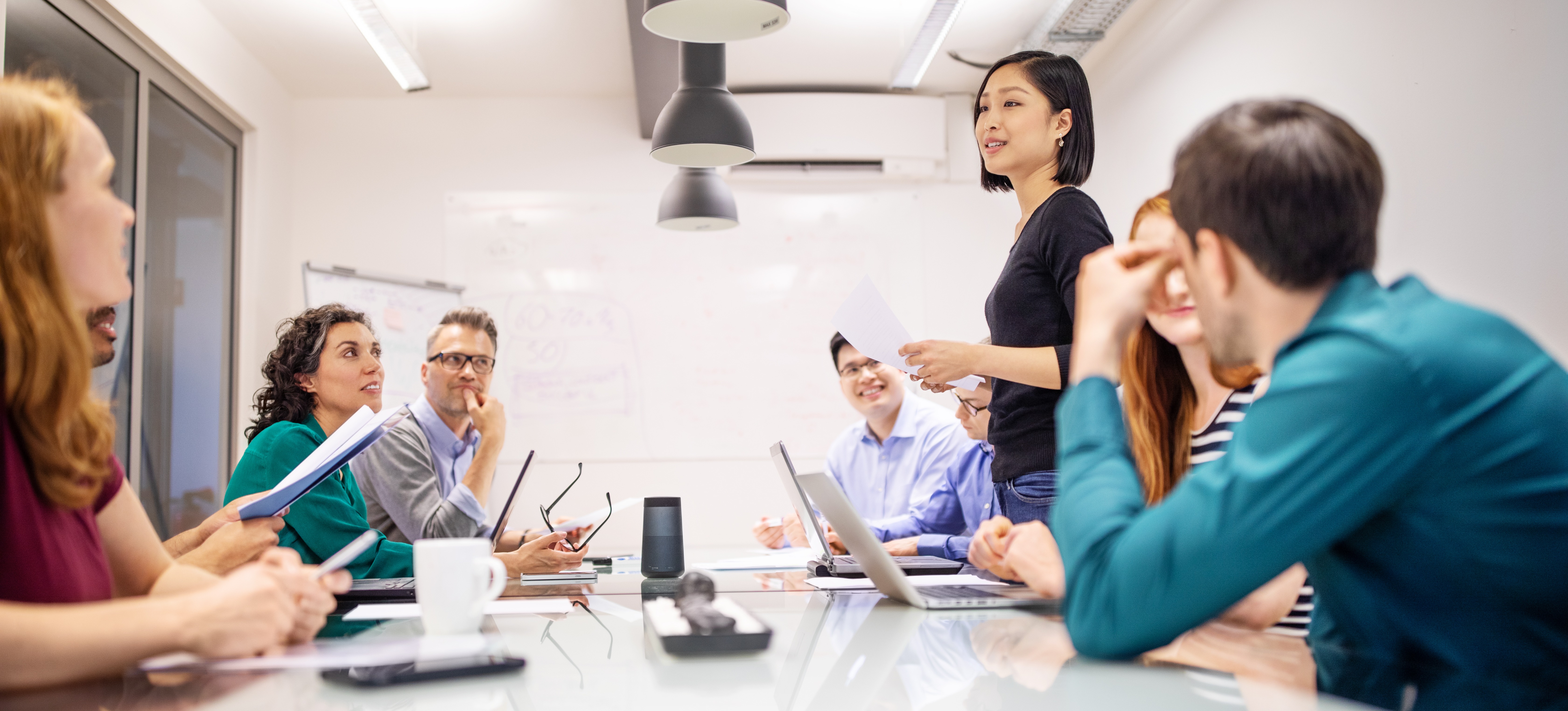 [Featured Image] A project team made up of multiple men and women sits in a meeting room to discuss a project that they're managing via a Kanban board.
