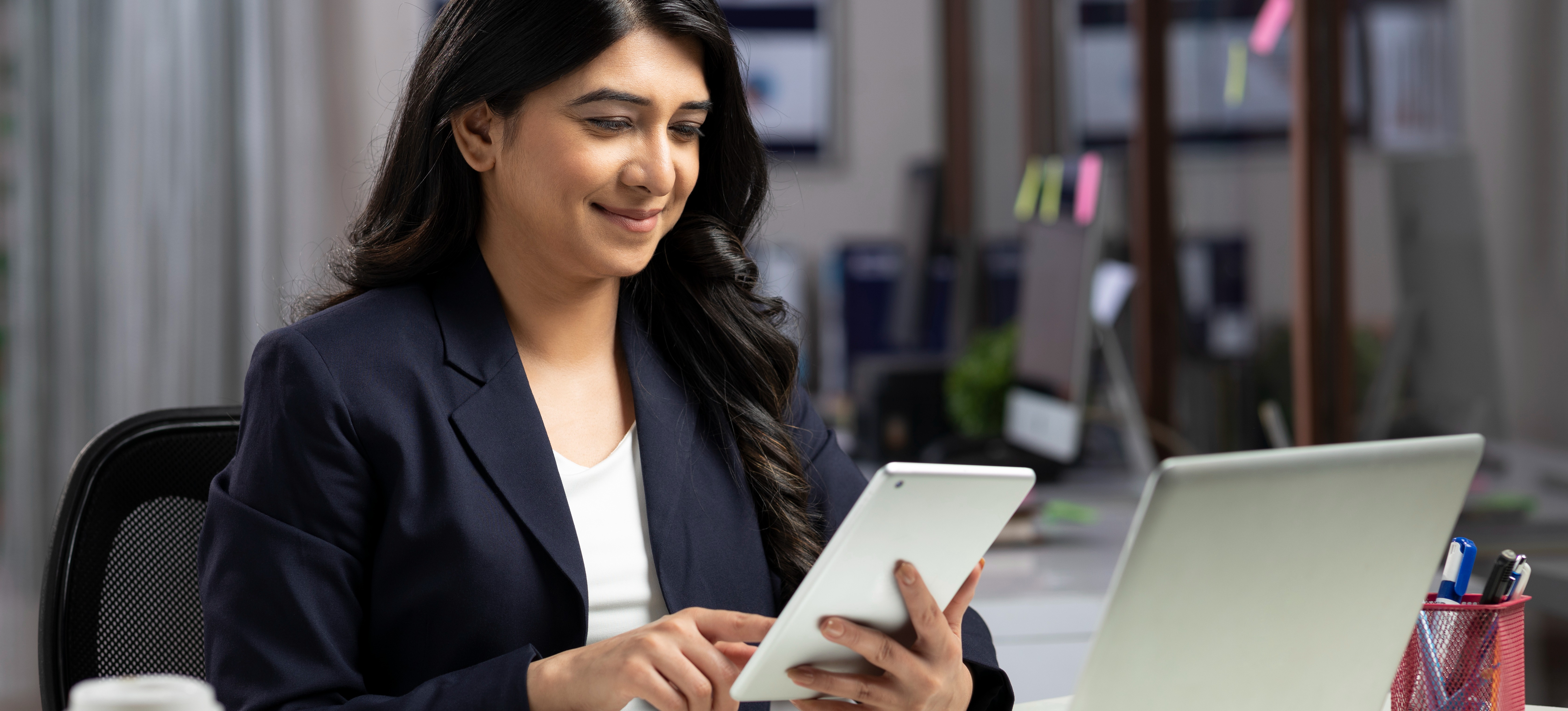 [Featured Image] A professionally dressed businessperson sitting in their office researches popular cybersecurity certifications on their tablet.