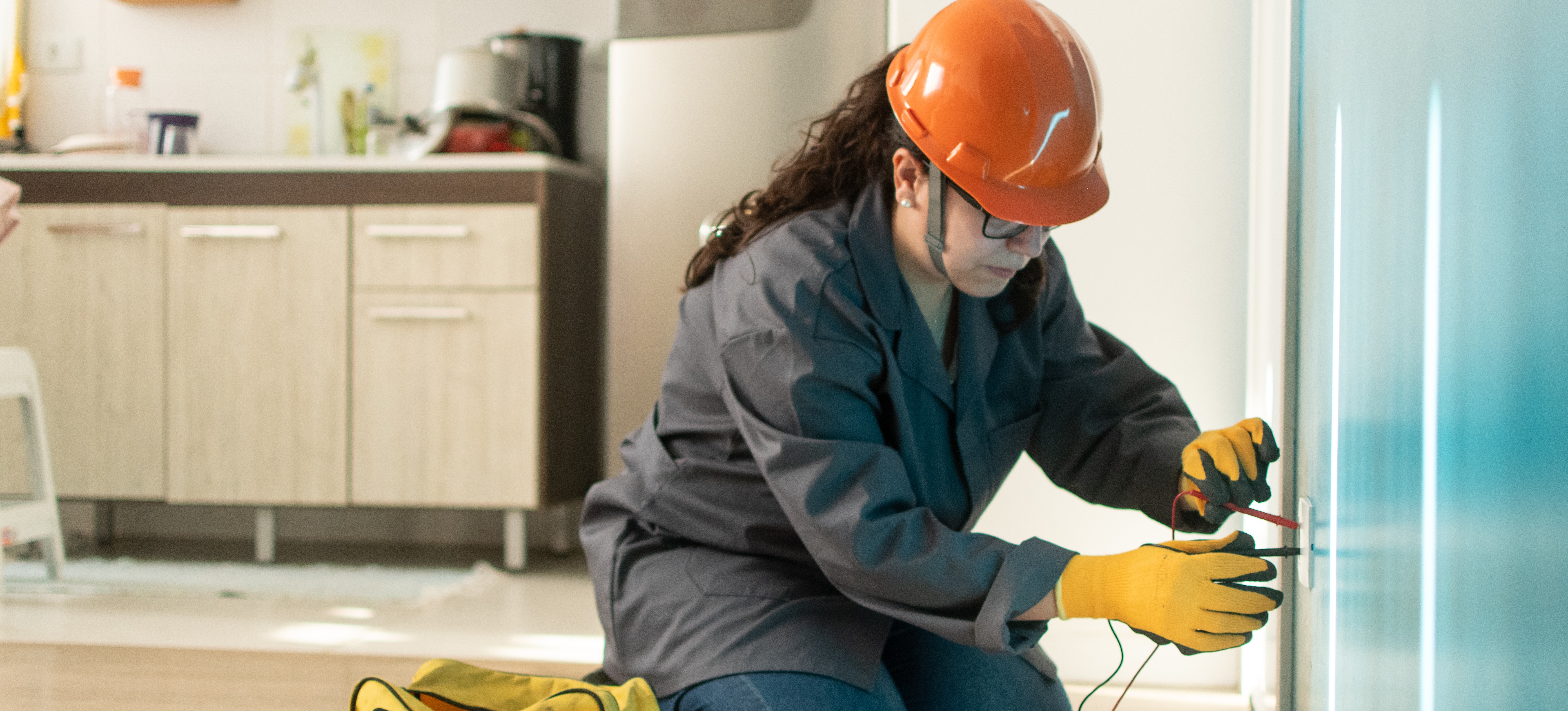 [Featured Image] A woman wearing a hard hat and gloves works as an electrician, one of the highest paying trade jobs currently available, using a multimeter to check for an electrical current in a private home.
