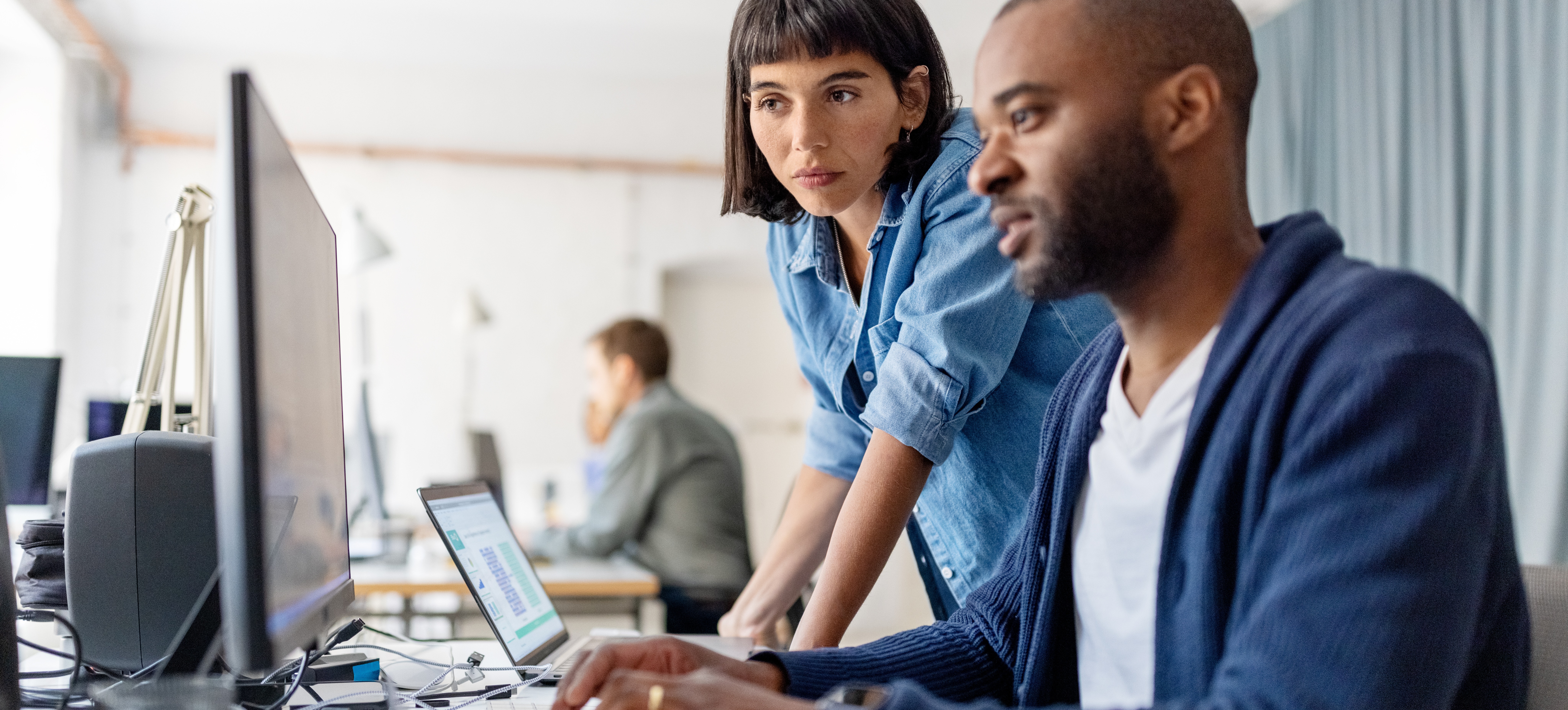 [Featured Image] Two coworkers discuss AI hallucinations on their computers in an open-space office.
