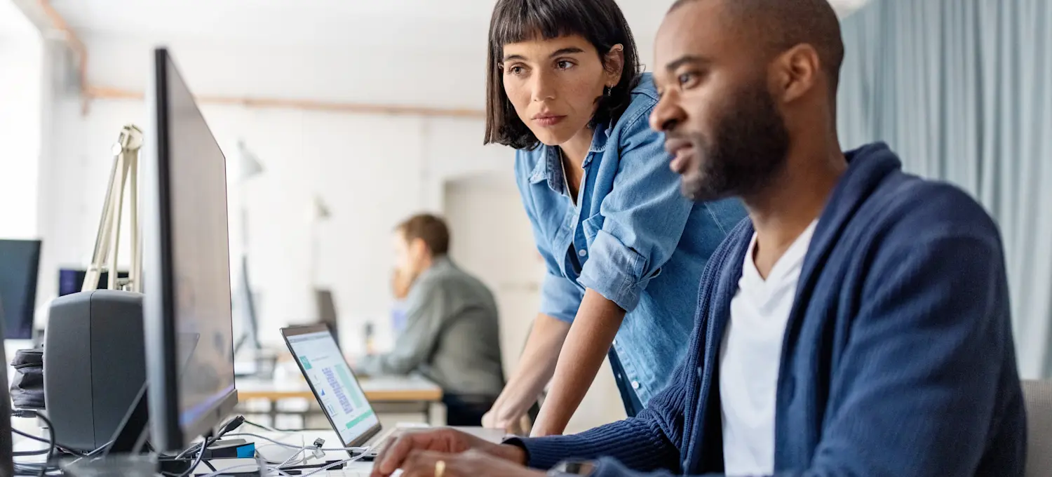 [Featured Image] Two coworkers discuss AI hallucinations on their computers in an open-space office.
