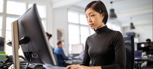 [Featured Image] A woman works at a desktop computer in an office.