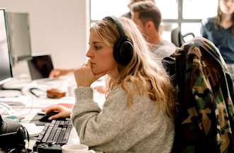 [Featured Image] A woman wearing headphones works on a desktop computer. 