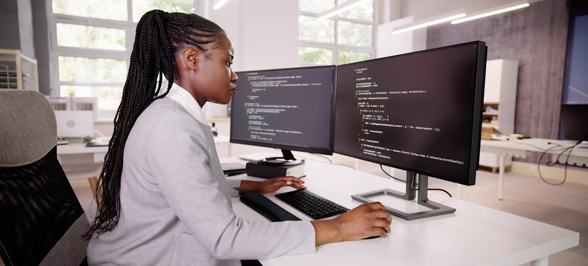 [Featured Image] In an empty office, a programmer in a gray jacket sits at a desktop computer with two screens and works on code using Objective-C.
