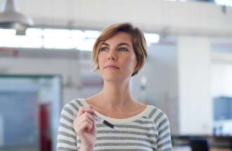 [Featured Image]:  A female, with short blonde hair, and wearing a white and gray striped top, is holding a pen in one hand and documents in the other hand. She is standing near her desk in her office. 