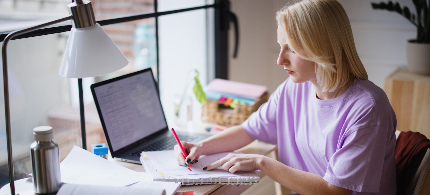 [Featured image] A blond student in a pink t-shirt sits at a desk writing in a notebook.