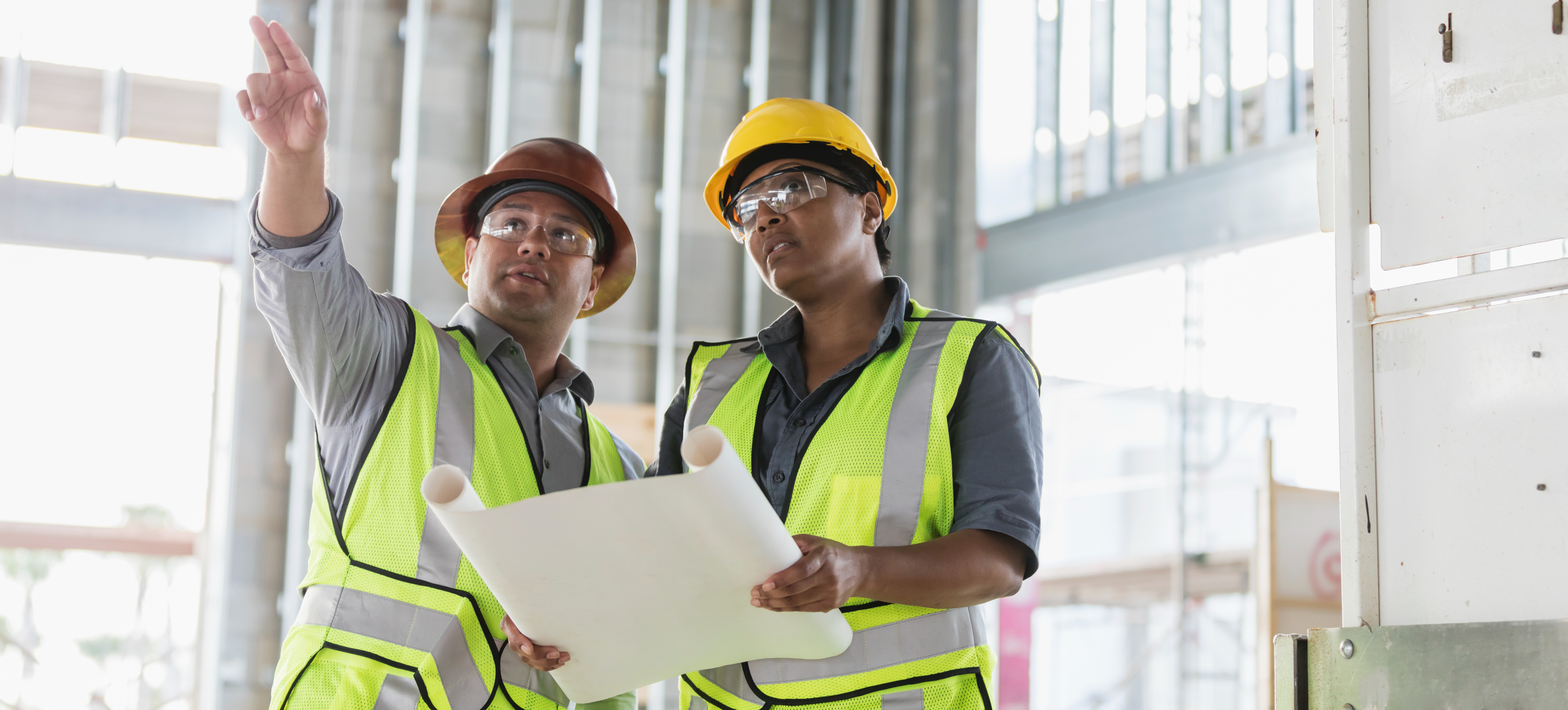 [Featured Image] Two construction project managers stand in the middle of a construction site in protective gear, one holding a blueprint and the other pointing to something off-camera.
