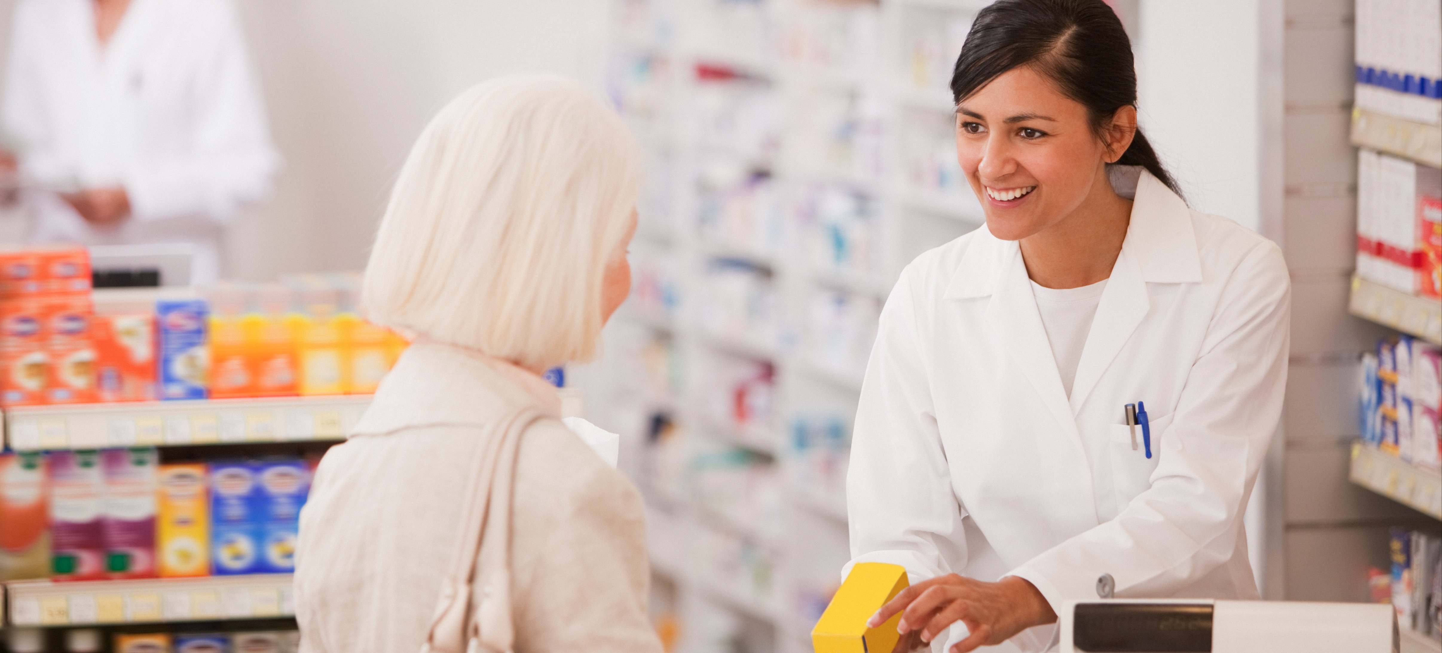 [Featured Image] A pharmacist who earned a PharmD degree answers a customer’s questions while holding a small yellow box over the pharmacy counter. 