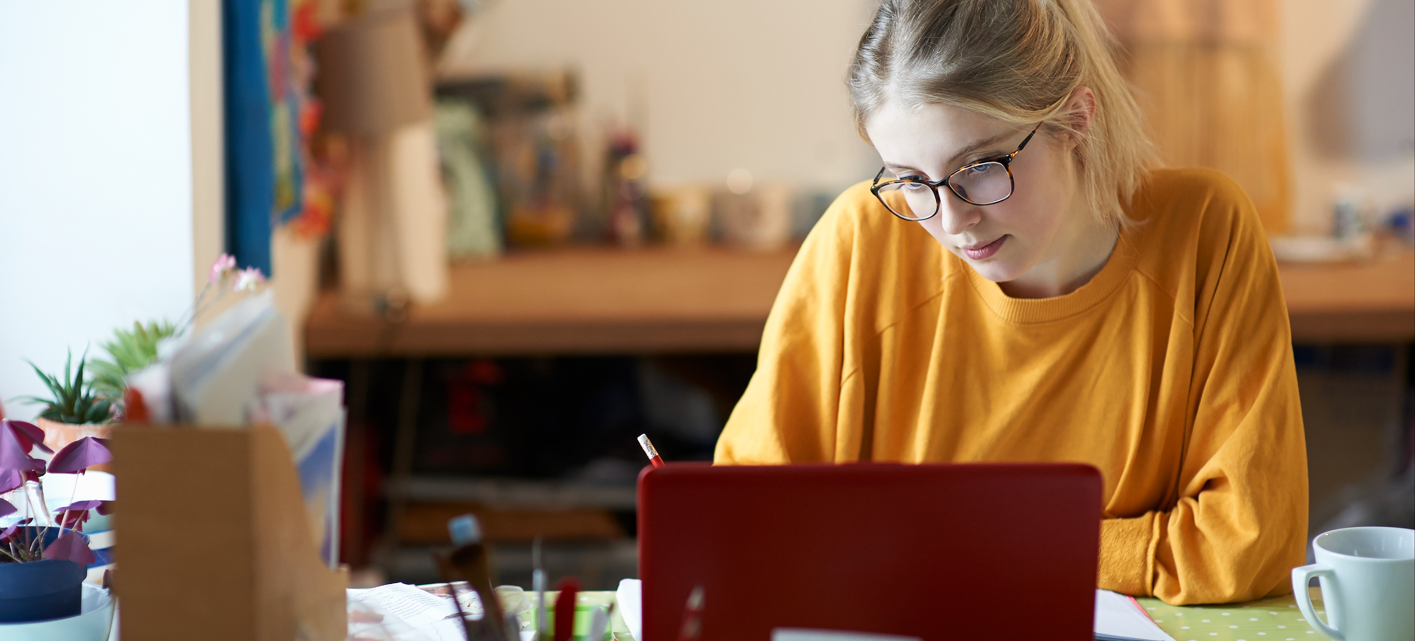 [FEATURED IMAGE] A college student wearing glasses studies at a desk with a laptop and pen in hand, pursuing a Bachelor of Fine Arts.