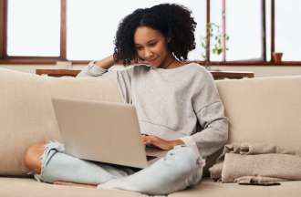 A young Black woman sits on a couch using her laptop. 