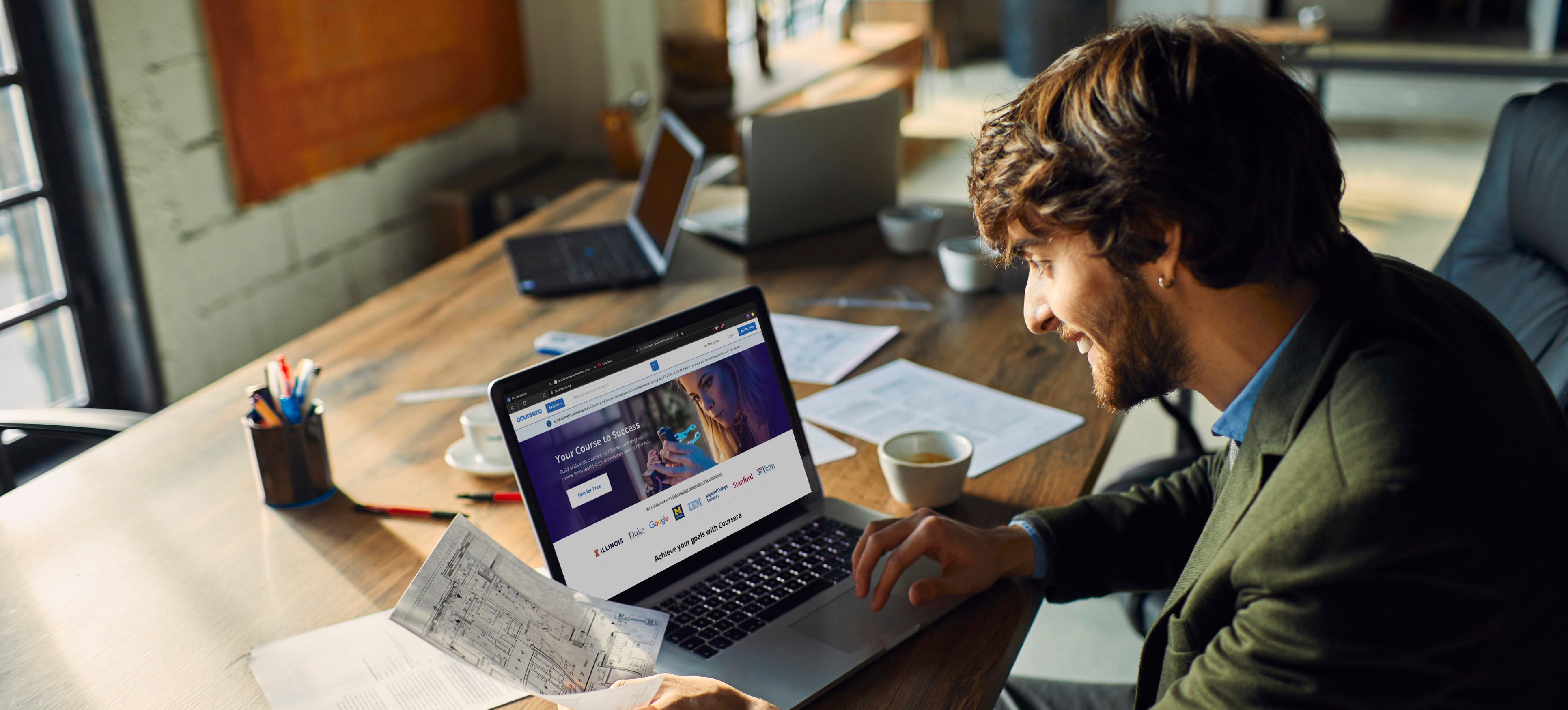 [Featured image] A marketing consultant wearing a blazer sits at a large table with a laptop and loose papers and reviews a website redesign for a client.
