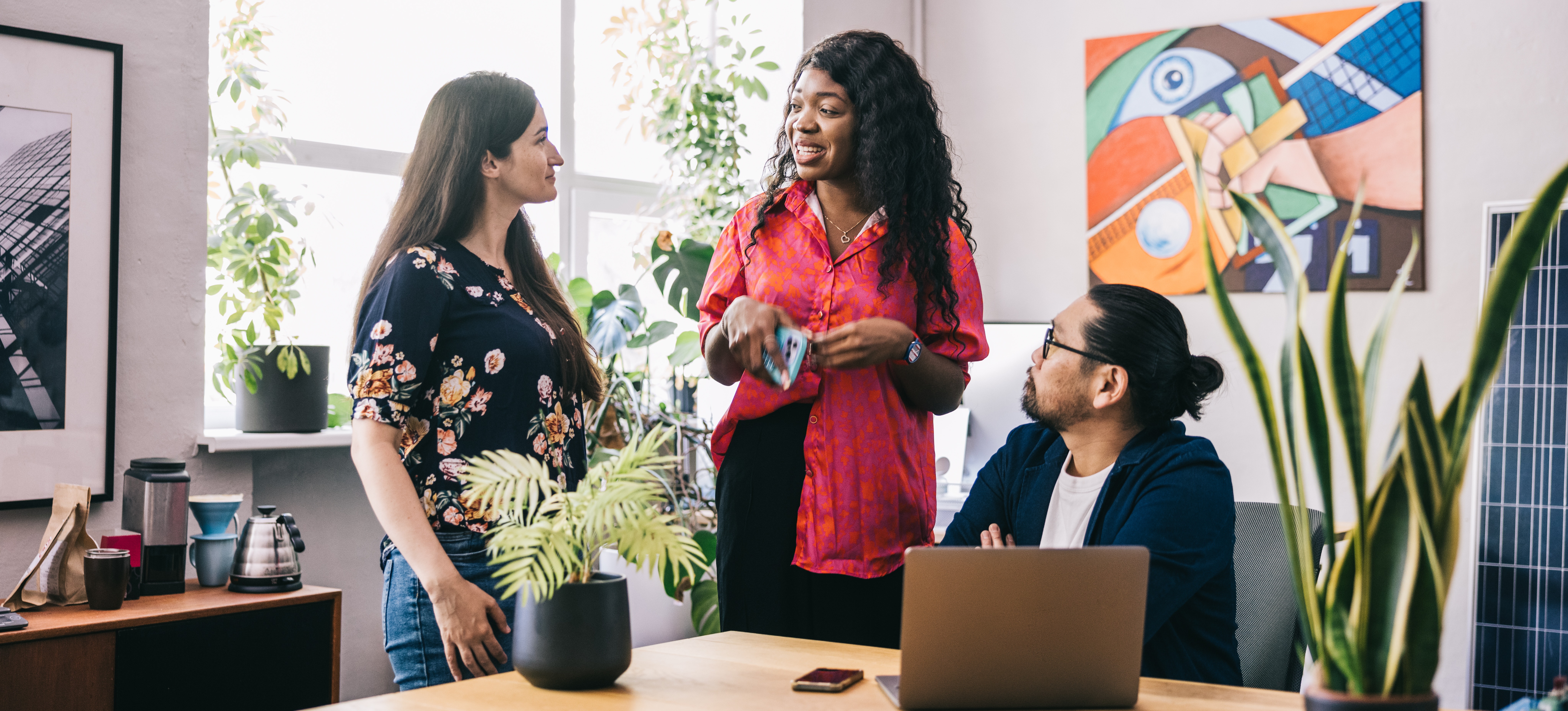 [Featured Image] A project manager and her team engage in conversation about continuous improvement as they stand in a sunny office.
