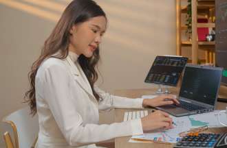 [Featured Image] A businesswoman is sitting at her desk with multiple monitors displaying graphs and trends created using DAX to analyze her data. 
