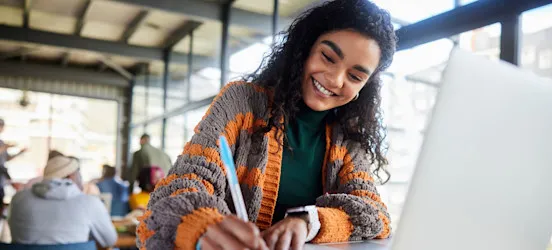 [Featured Image] A college student sitting with a laptop in the campus commons area working on their cover letter for graduate school.

