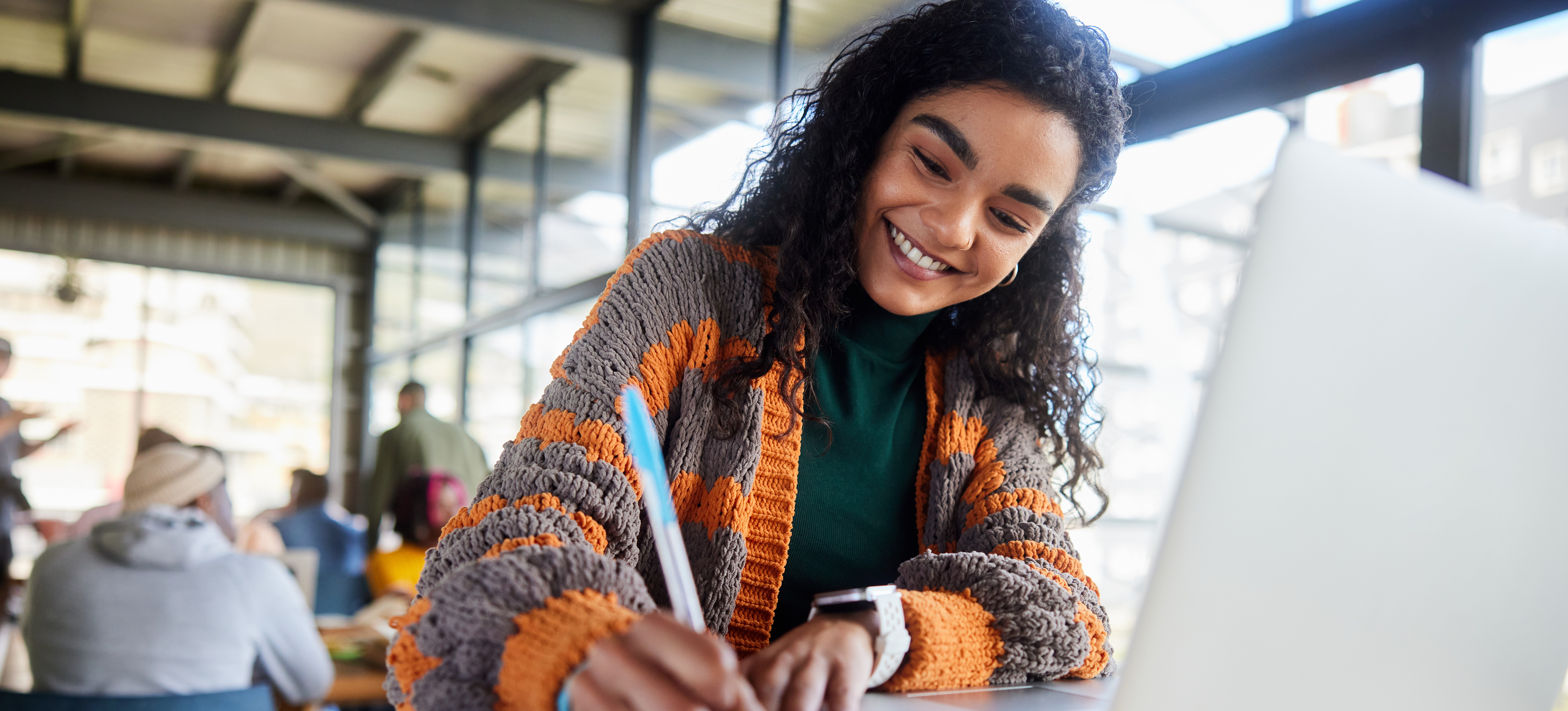 [Featured Image] A college student sitting with a laptop in the campus commons area working on their cover letter for graduate school.

