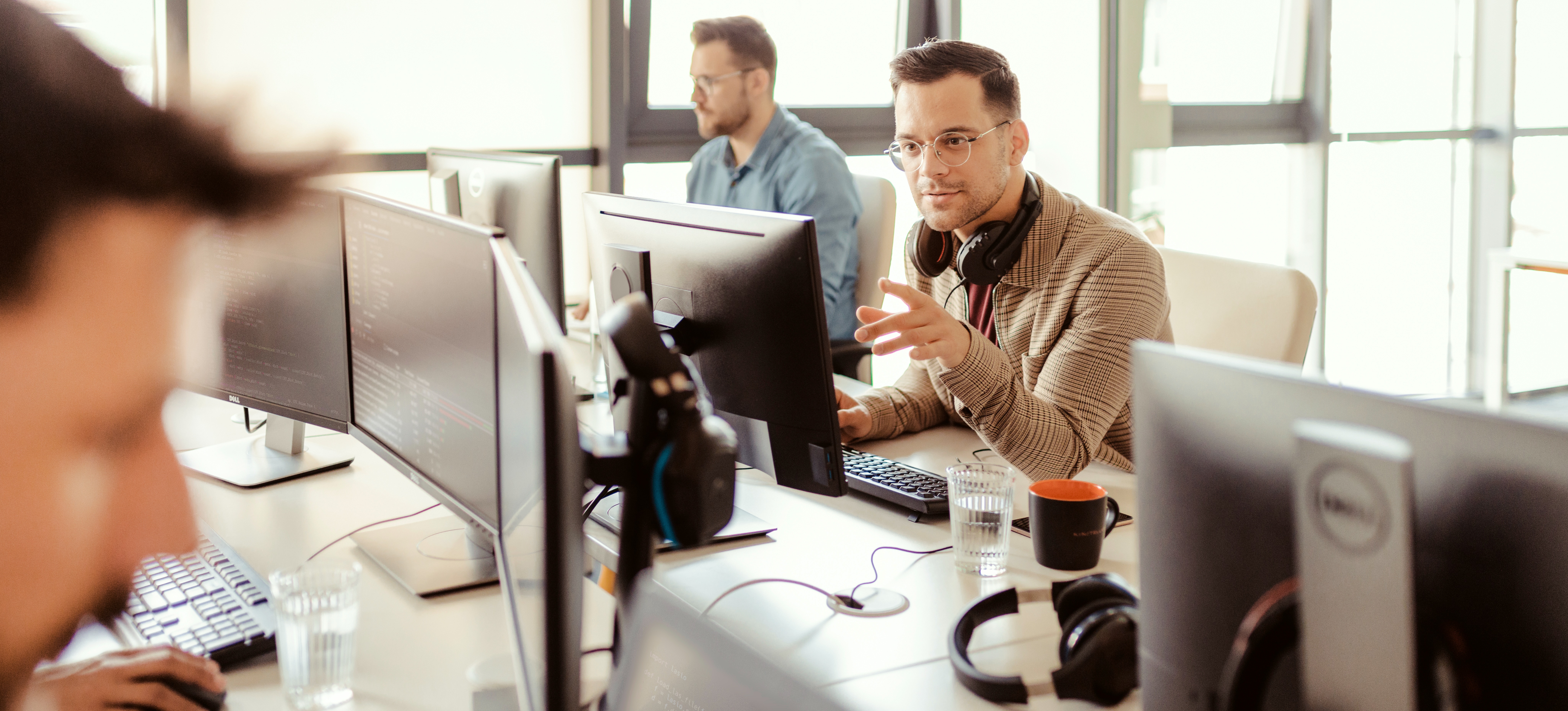 [Featured Image] A cybersecurity professional sits at his office shared space with others, working on his computer using MITRE ATT&CK to help prevent security breaches.
