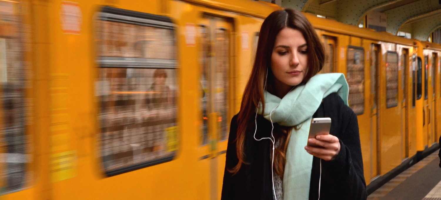 [Featured image] A student who attends a liberal arts college, wearing a black jacket and a light blue scarf, stands in front of a yellow train looking at their phone. 