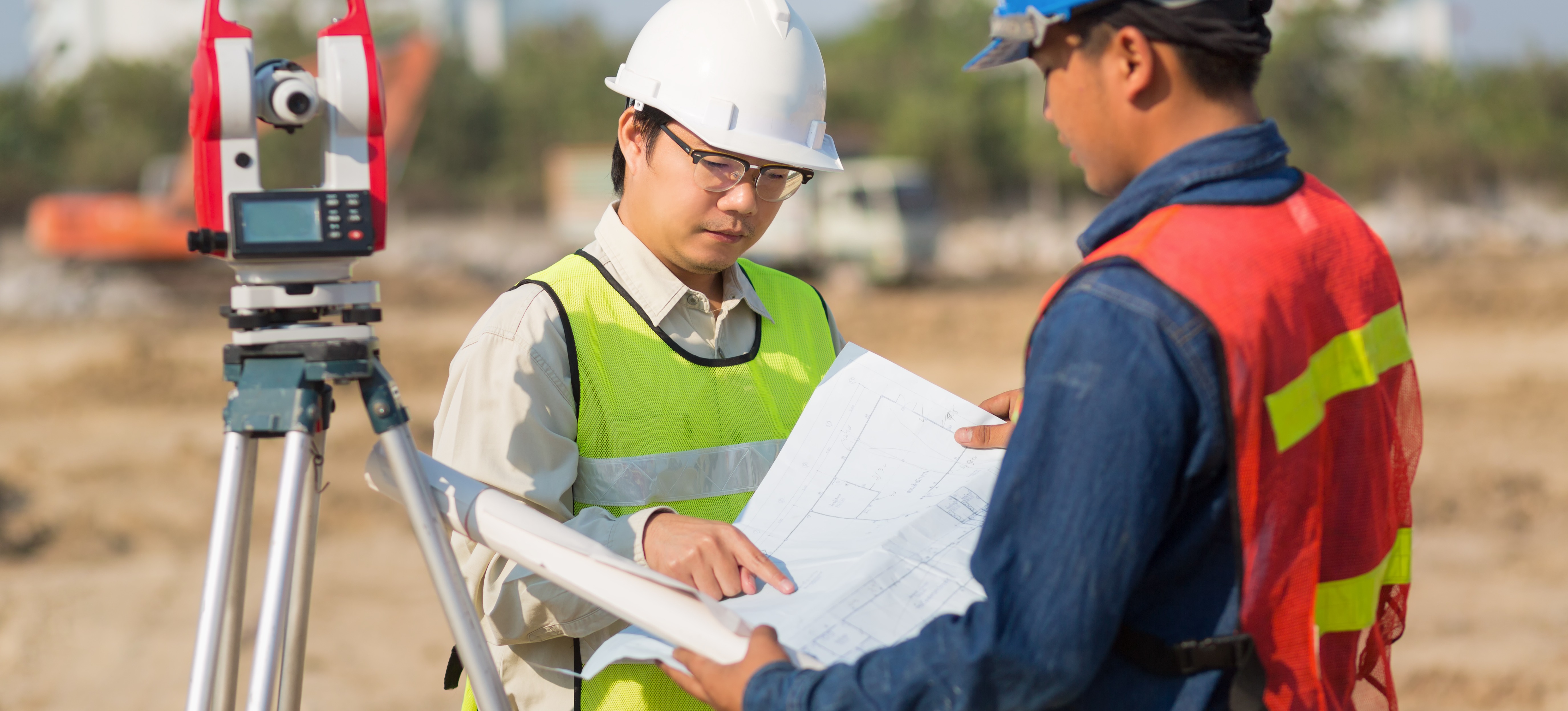 [Featured Image] Two engineers wearing hard hats and reflective vests stand outside at a job site looking at project plans.