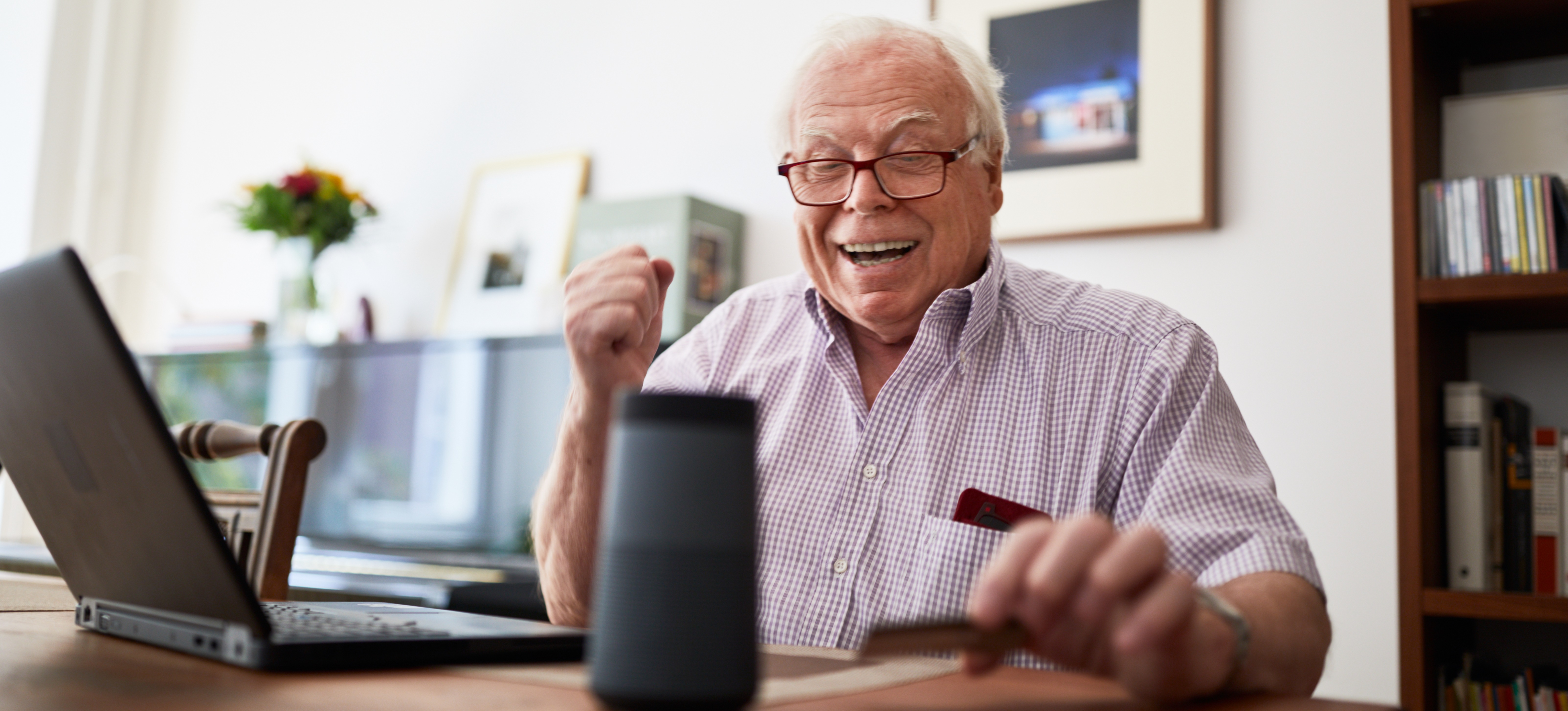 [Featured Image] An older person wearing glasses while sitting at a desk in front of an open laptop smiles as they use a natural language processing application example: a voice-activated assistant. 
