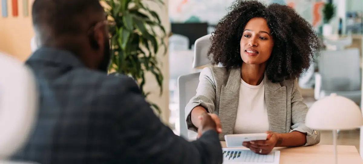 [Featured Image] A recruiter who uses ChatGPT to streamline hiring processes shakes hands with a new hire after an interview as they sit across the table from one another.