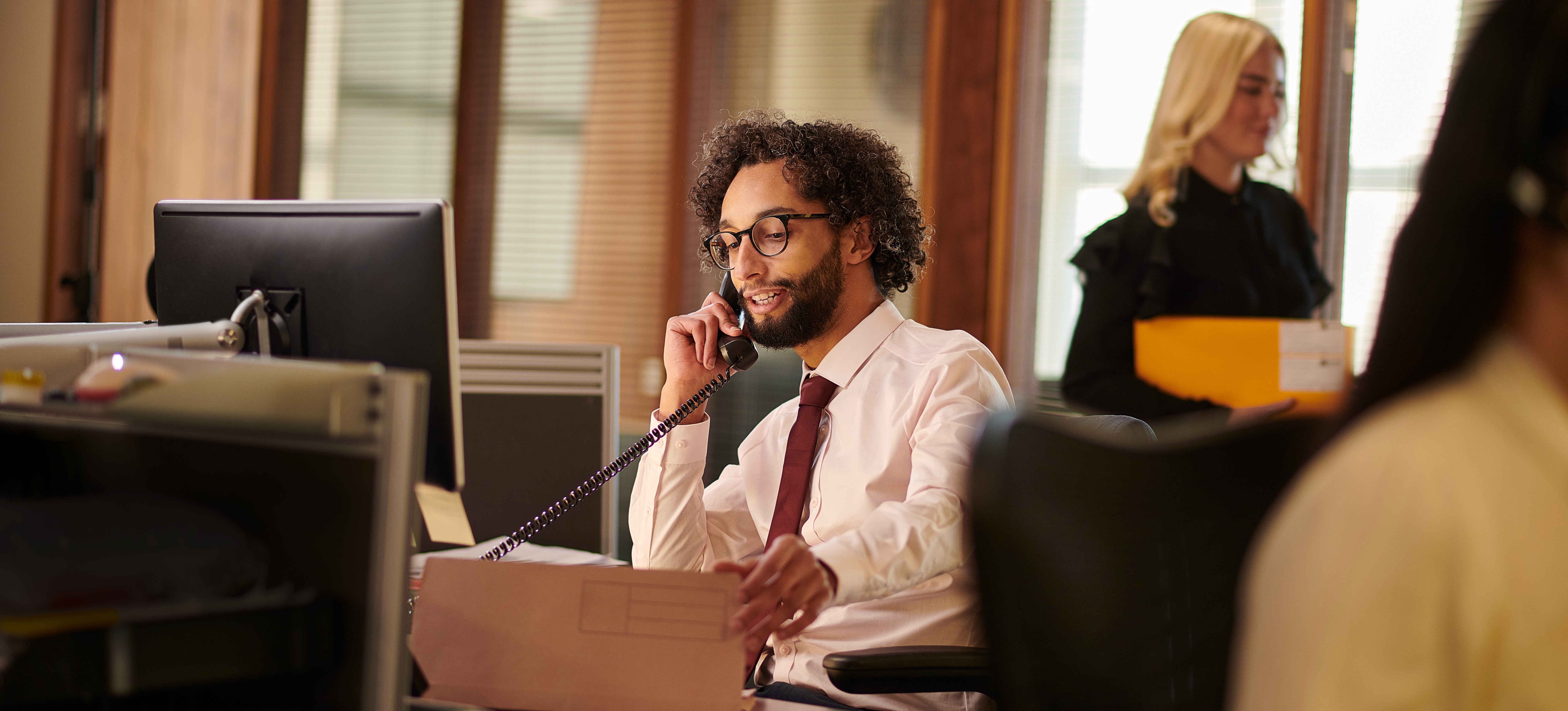 [Featured Image] A paralegal works at their desk in a busy office, talking to someone on the phone as they look through files.
