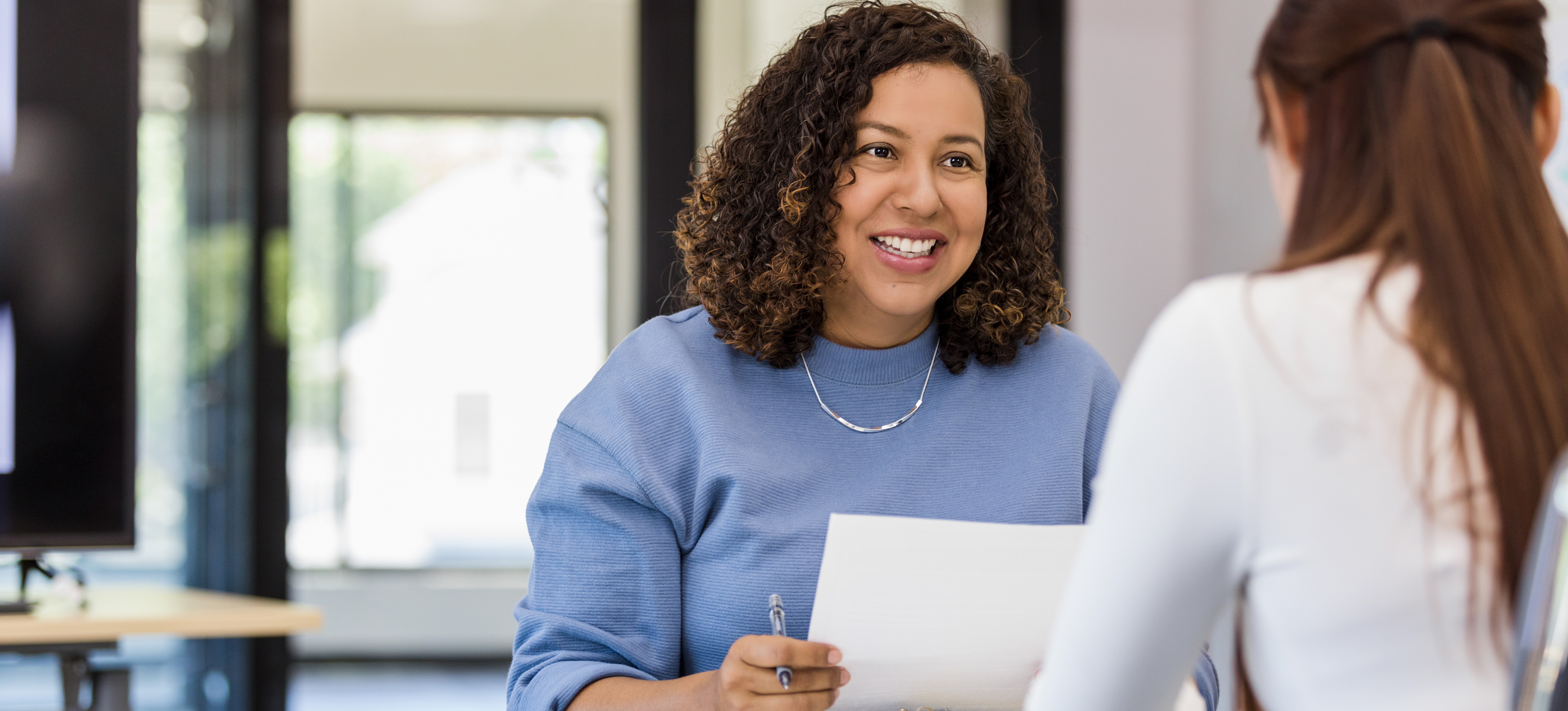 [Featured Image] A smiling employer sits at a table holding a piece of paper and pen and asks a job candidate software engineer interview questions.
