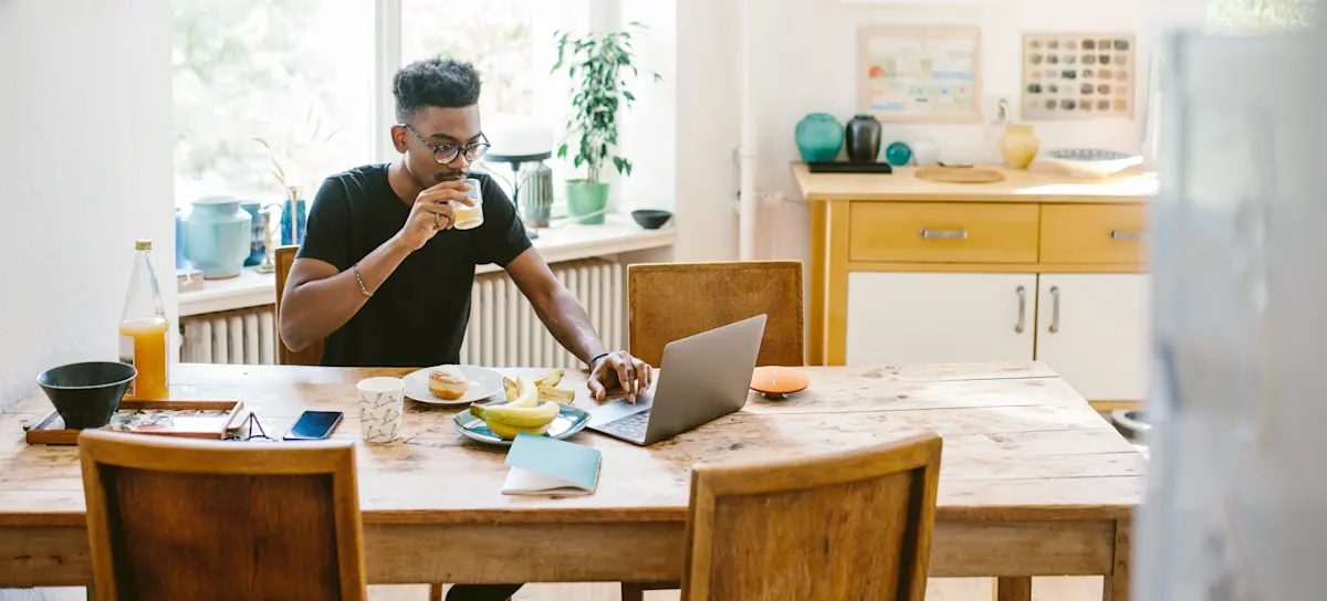 [Featured Image] A person drinking orange juice uses their laptop at home on their dining room table.
