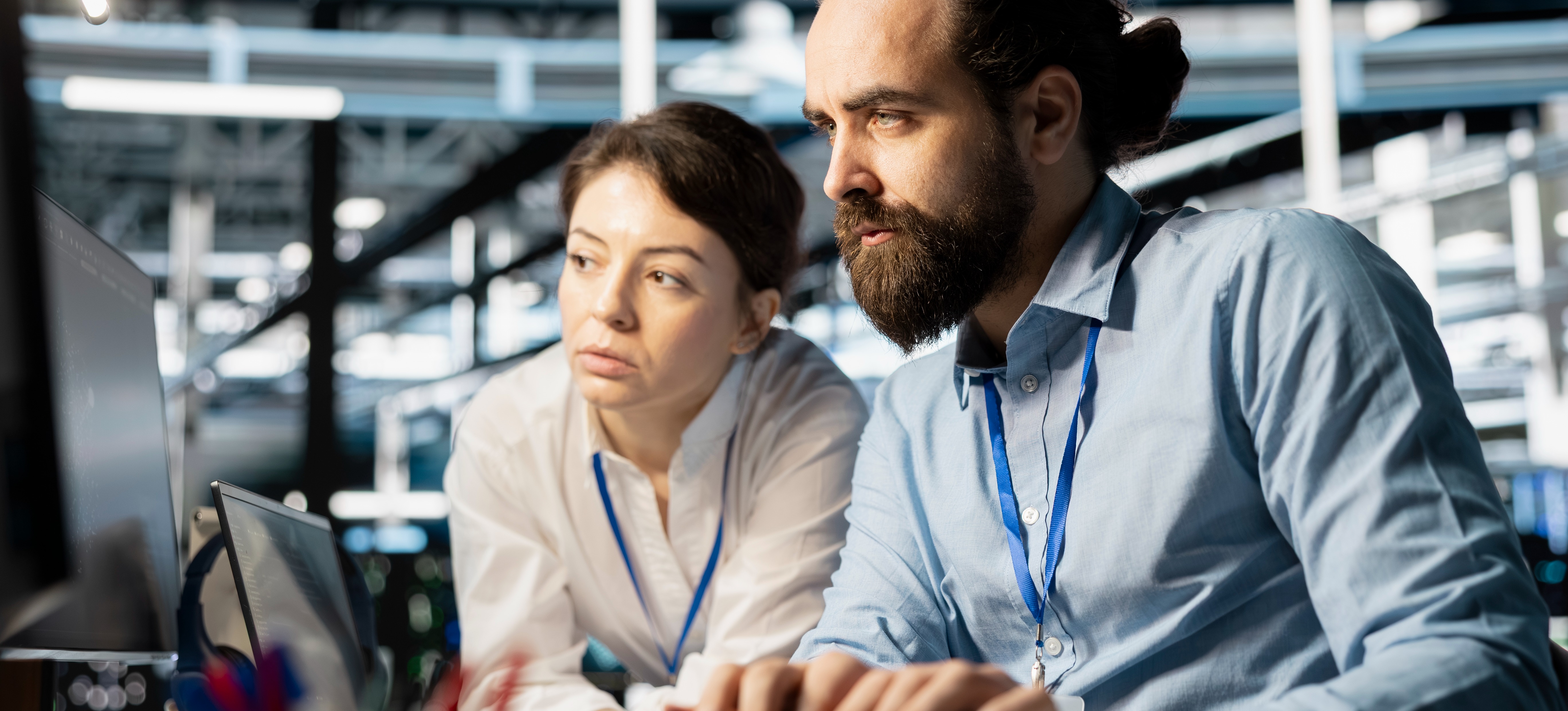 [Featured Image] Two machine learning professionals use neural architecture search as they both look at a computer screen in a large office space.

