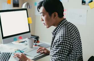 [Featured Image] A person in a checkered button-down shirt works with R programming at a desk with a laptop, monitor, and keyboard. There are colored Post-Its hanging on the monitor and wall to the right.