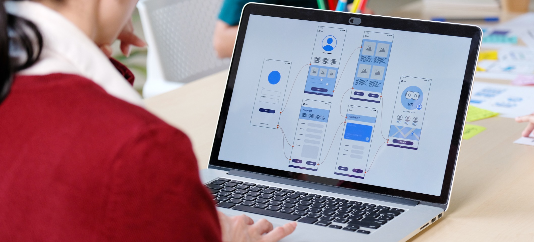 [ Image en vedette ] Un designer UX vêtu d'un pull rouge travaille sur les schémas de câblage d'un prototype haute fidélité sur son ordinateur portable, dans un bureau partagé.