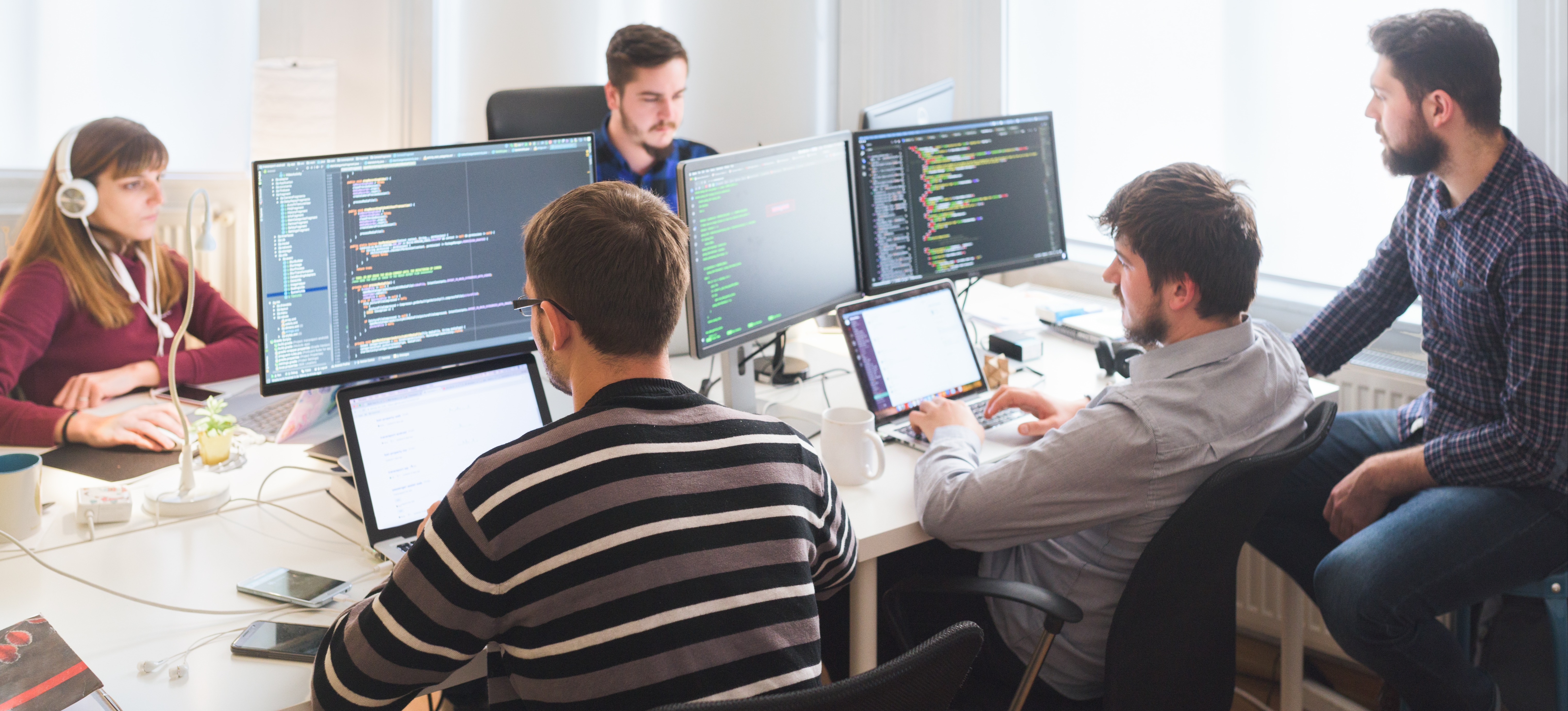 [Featured Image] A team of four men and one women sit at a desk, each at their own computer, focusing on their software engineering projects in the office.