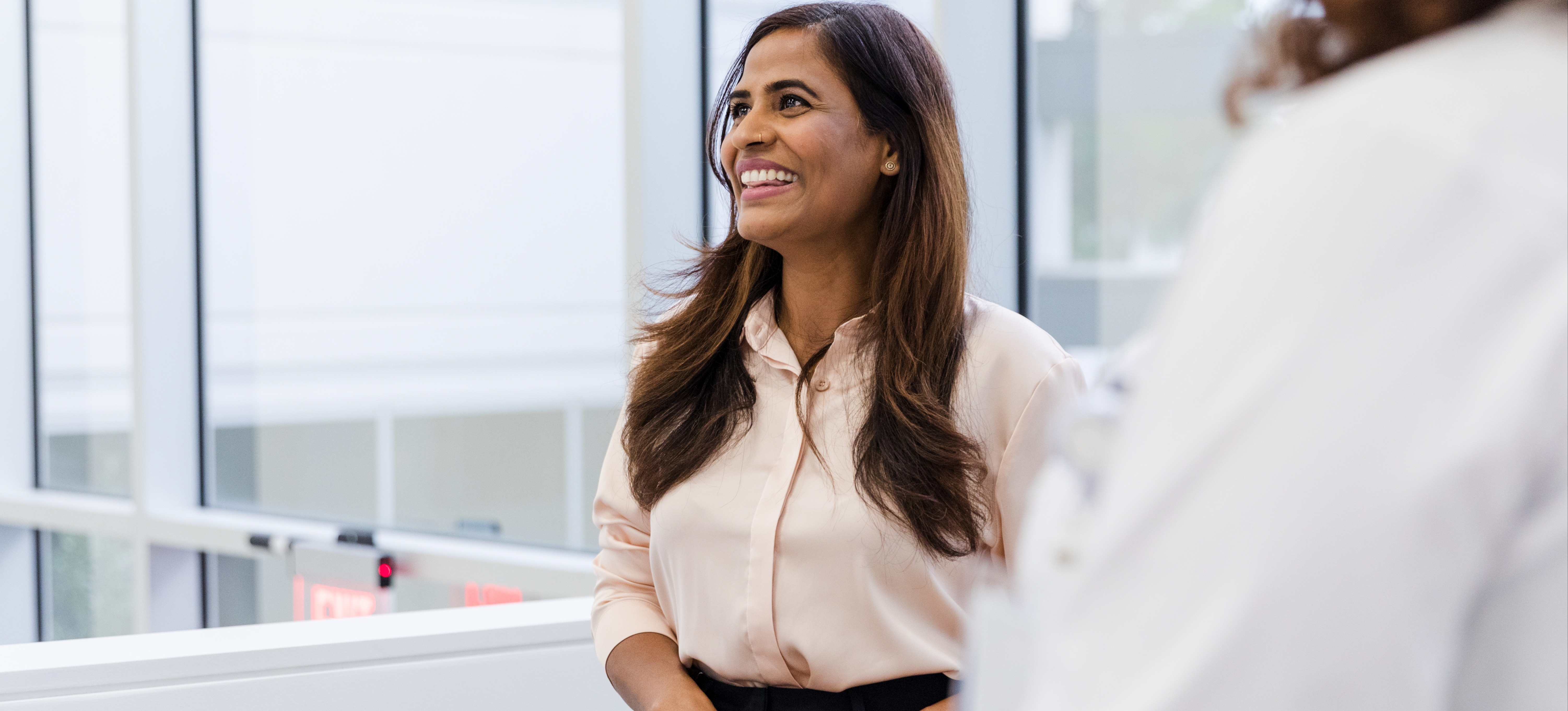 [Featured Image] I smiling coworker stands in a windowed office and speaks with their colleagues about the types of MBAs to pursue.
