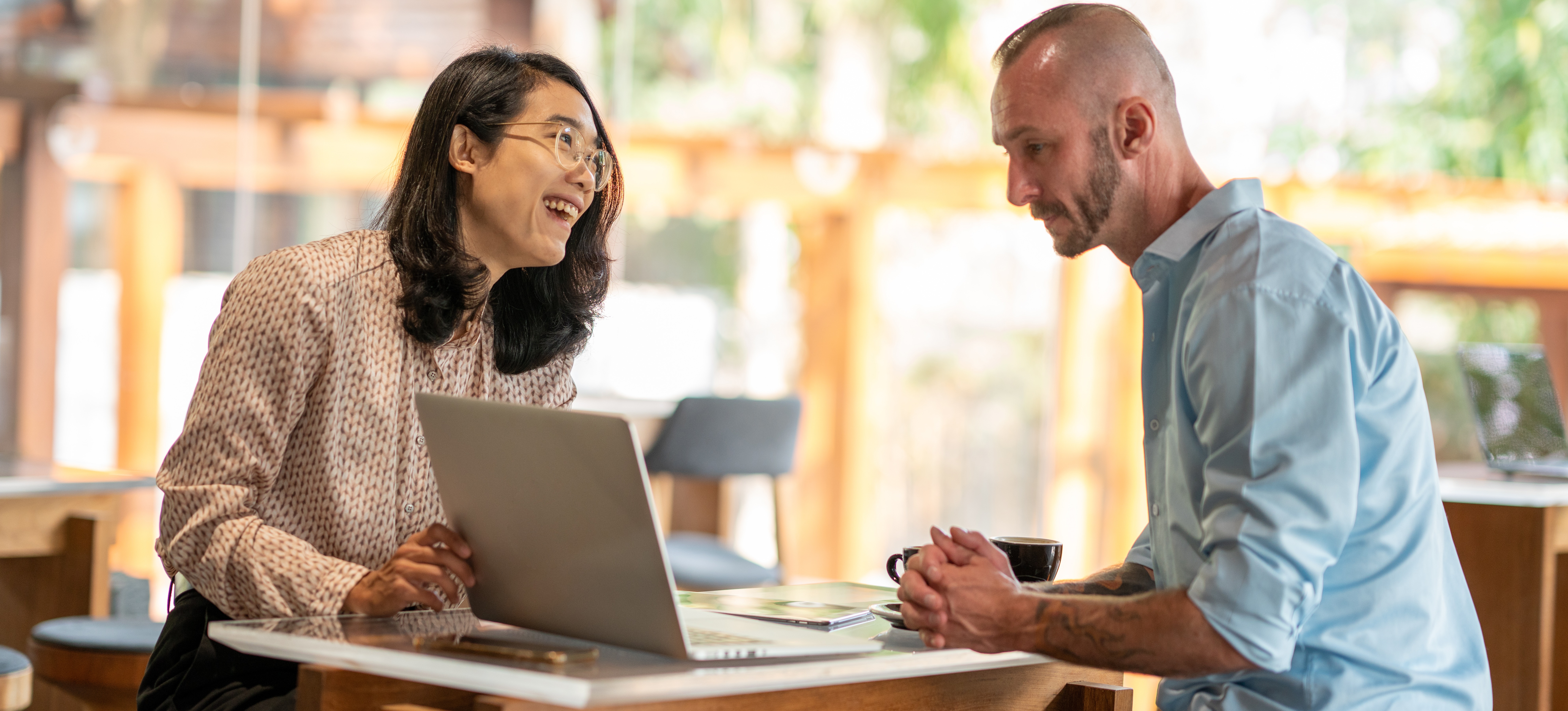 [Featured Image] Two coworkers sit at a workspace, talking and examining a laptop screen.
