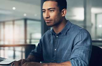 [Featured image] A person wearing a blue button-up shirt with the sleeves rolled up to the elbows sits in front of their desktop computer. 