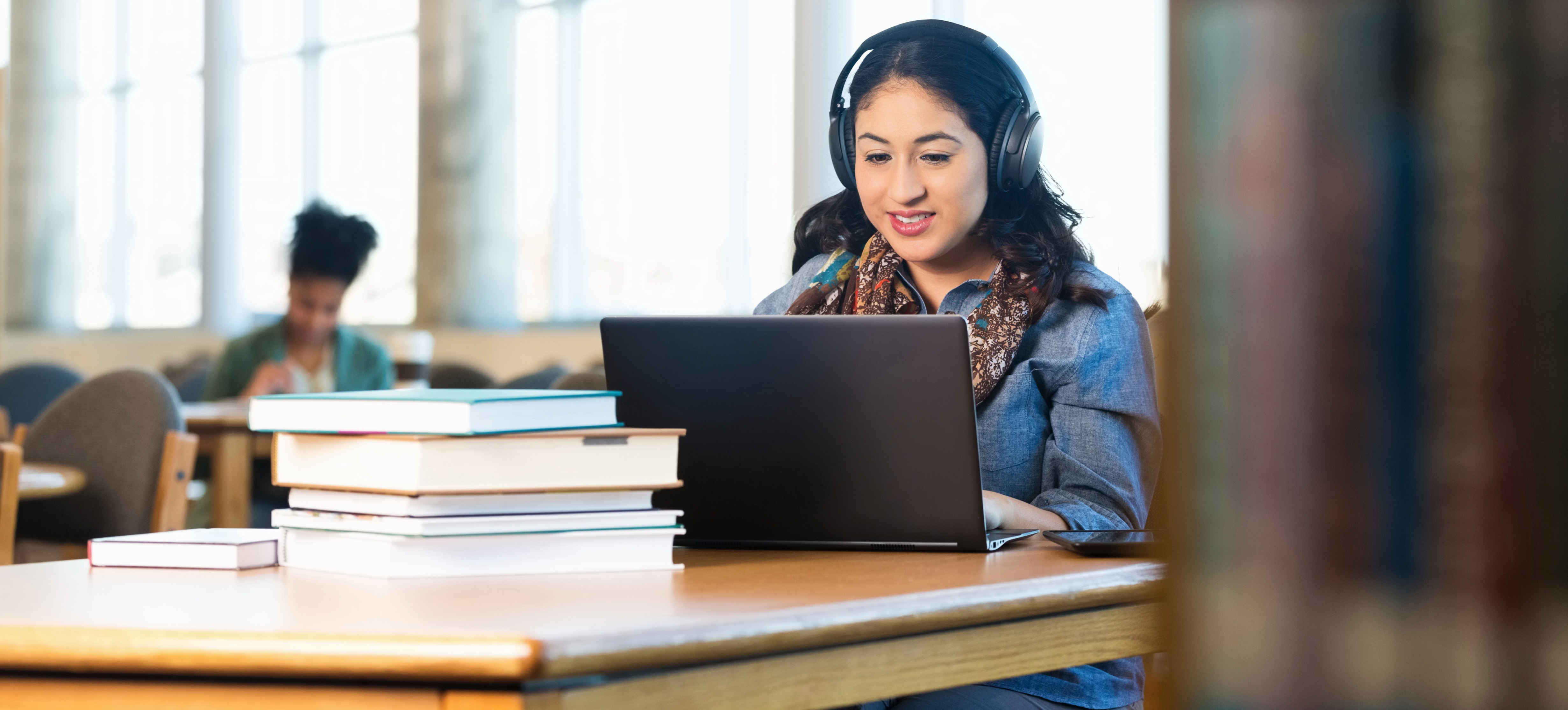 [Featured image] College student studying with a laptop and books at a table in a library, smiling and wearing headphones.