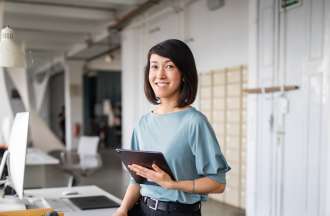 [Featured image] A female data analyst, wearing a gray short sleeve shirt, is standing in front of her desk, holding a laptop. The desk has a desktop computer and other supplies on it.