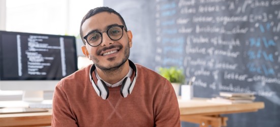 [Featured image] A data scientist in a sweater and glasses sits in front of a computer with programming language on the screen.