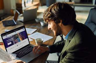 [Featured image] A marketing consultant wearing a blazer sits at a large table with a laptop and loose papers and reviews a website redesign for a client.