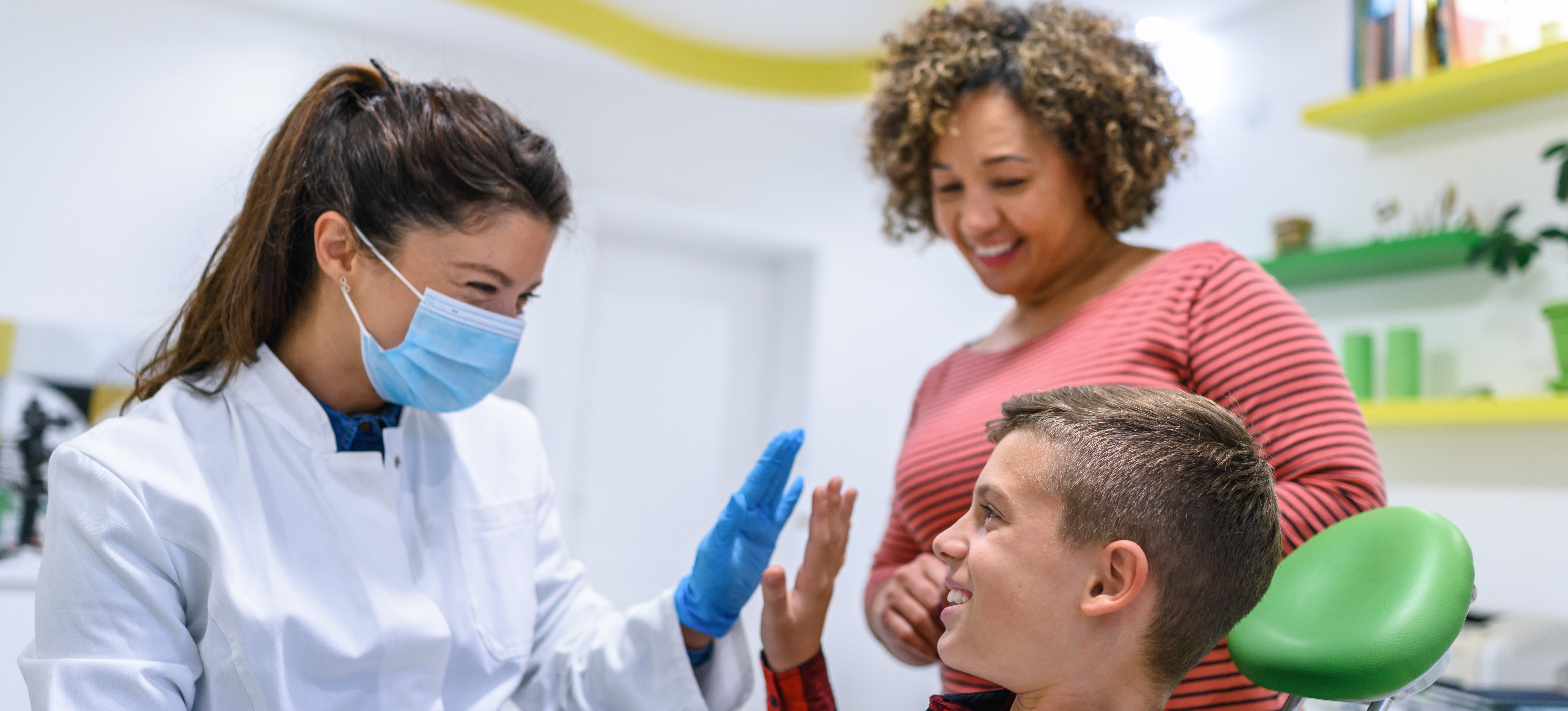 [Featured Image] An orthodontist, which is a low-stress job, high-fives a patient in the dental chair while their parent looks on. 
