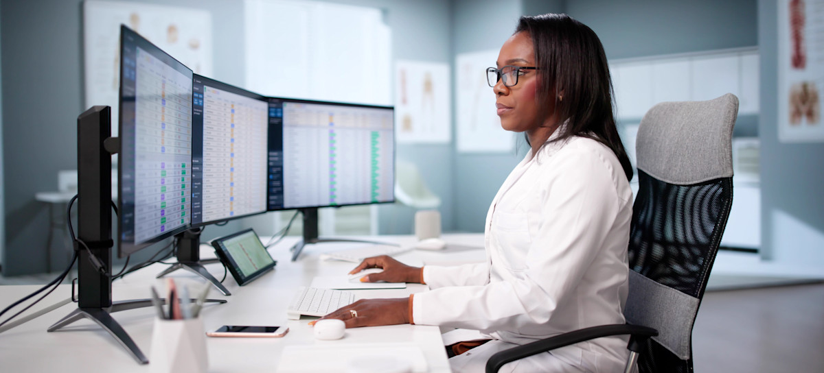 [Featured Image] A doctor dressed in white sits at their clinic desk and views spreadsheets on three monitors, working on converting Excel columns to rows.
