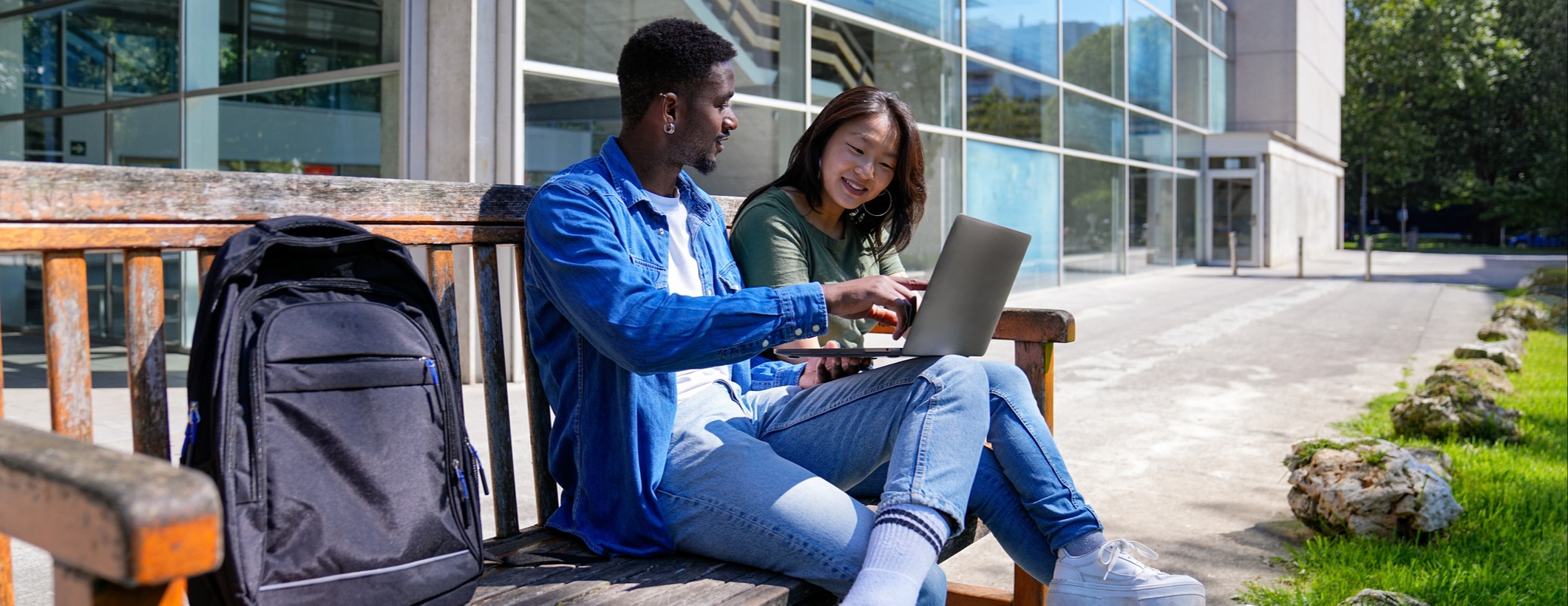 [Featured Image] Two learners sit on a bench outside discussing data mining architecture and reviewing their notes from class on a laptop computer.
