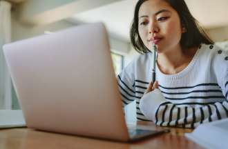 A young woman holding a pen to her lips ponders information she's reading on a laptop. 