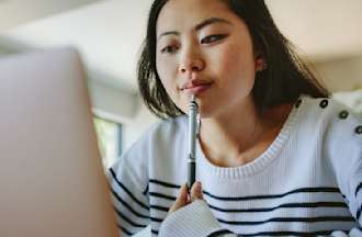 A young woman holding a pen to her lips ponders information she's reading on a laptop.