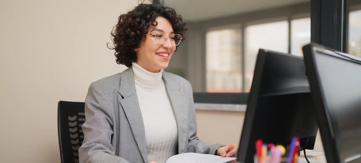 [Featured Image] A smiling content creator working for a marketing firm uses their content optimization skills as they work at their computer.
