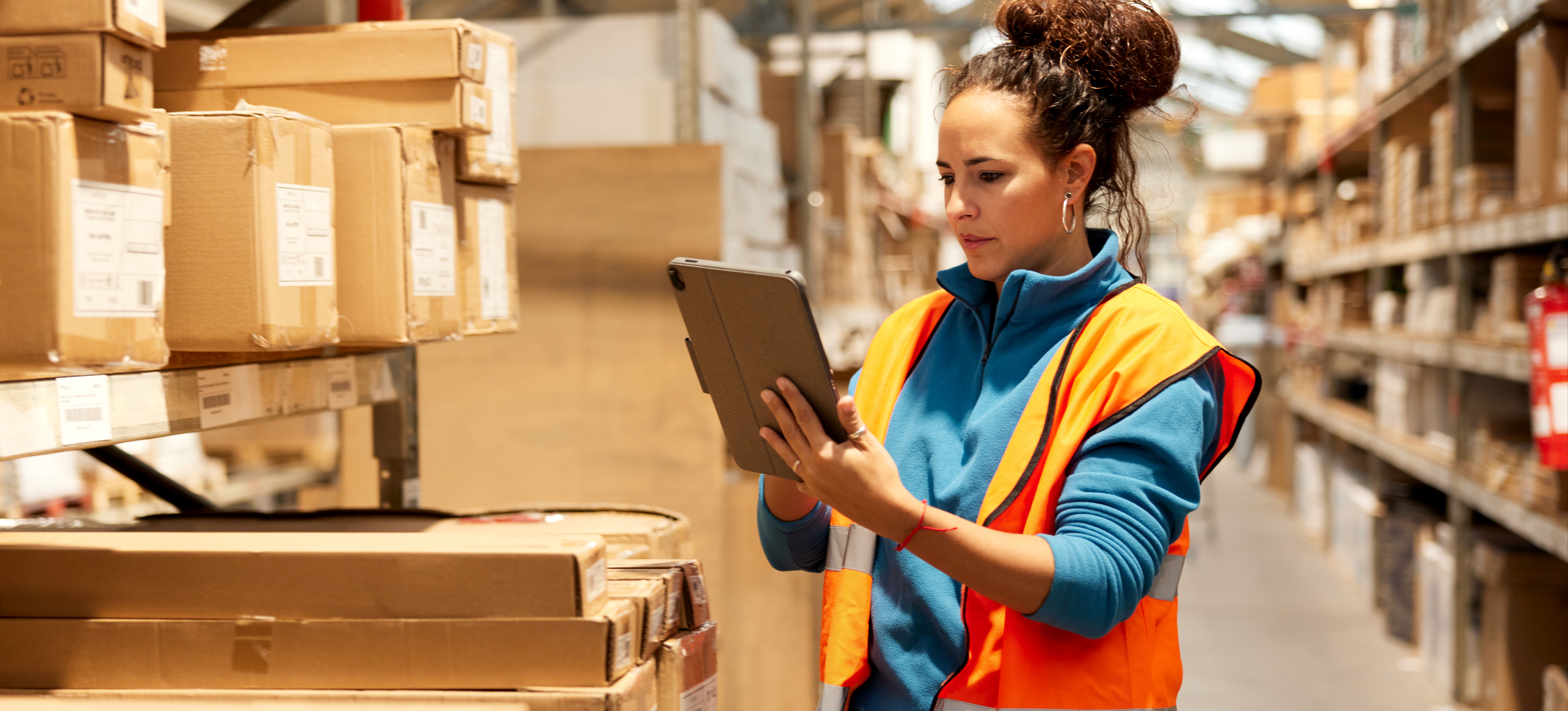 [Featured Image] Wearing an orange safety vest, a person working for an inventory control analyst salary examines information on their tablet inside a stocked warehouse.
