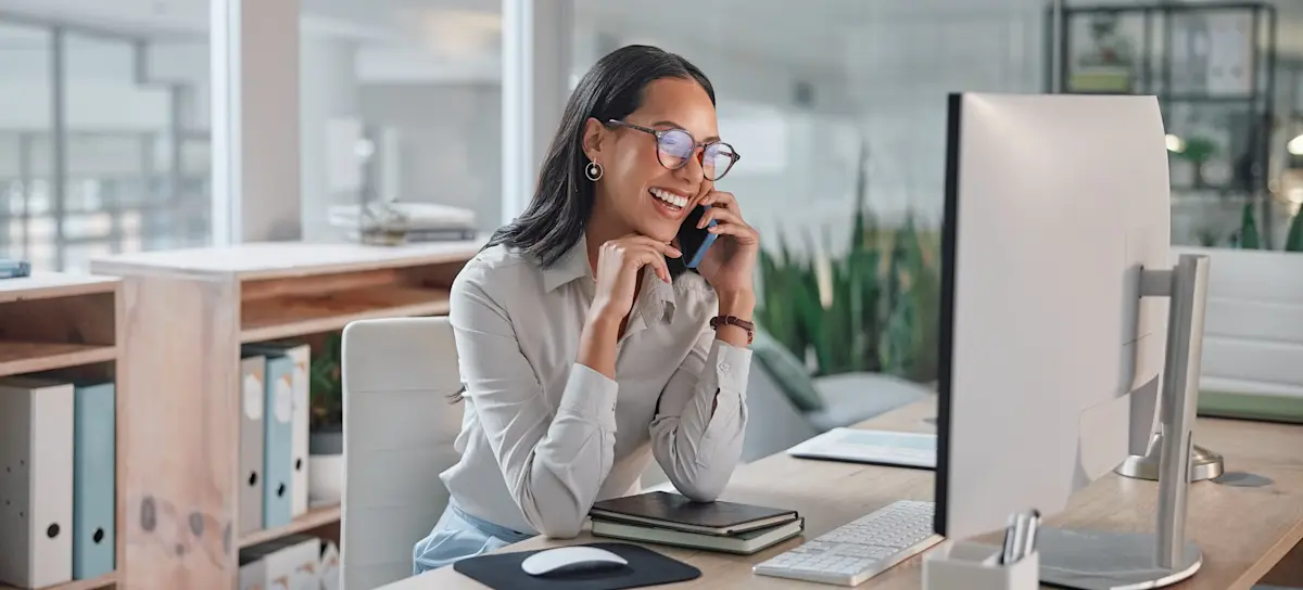 [Featured Image] A person working for a business development associate salary sits at their desk and laughs on the phone while looking at their desktop computer.

