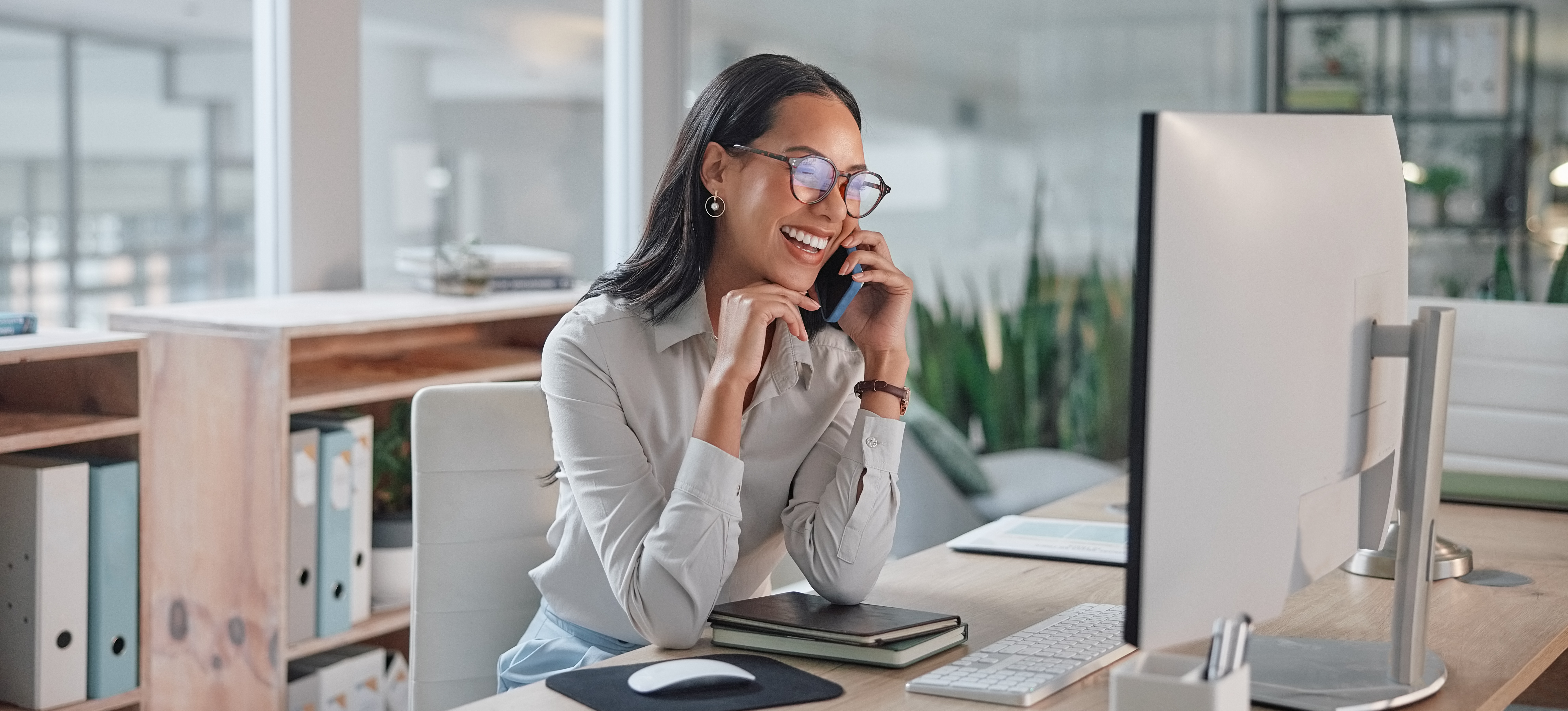 [Hauptbild] Eine Frau, die für den Gehaltsscheck einer Business Development Associate arbeitet, sitzt an ihrem Schreibtisch und lacht am Telefon, während sie auf ihren Desktop-Computer schaut.