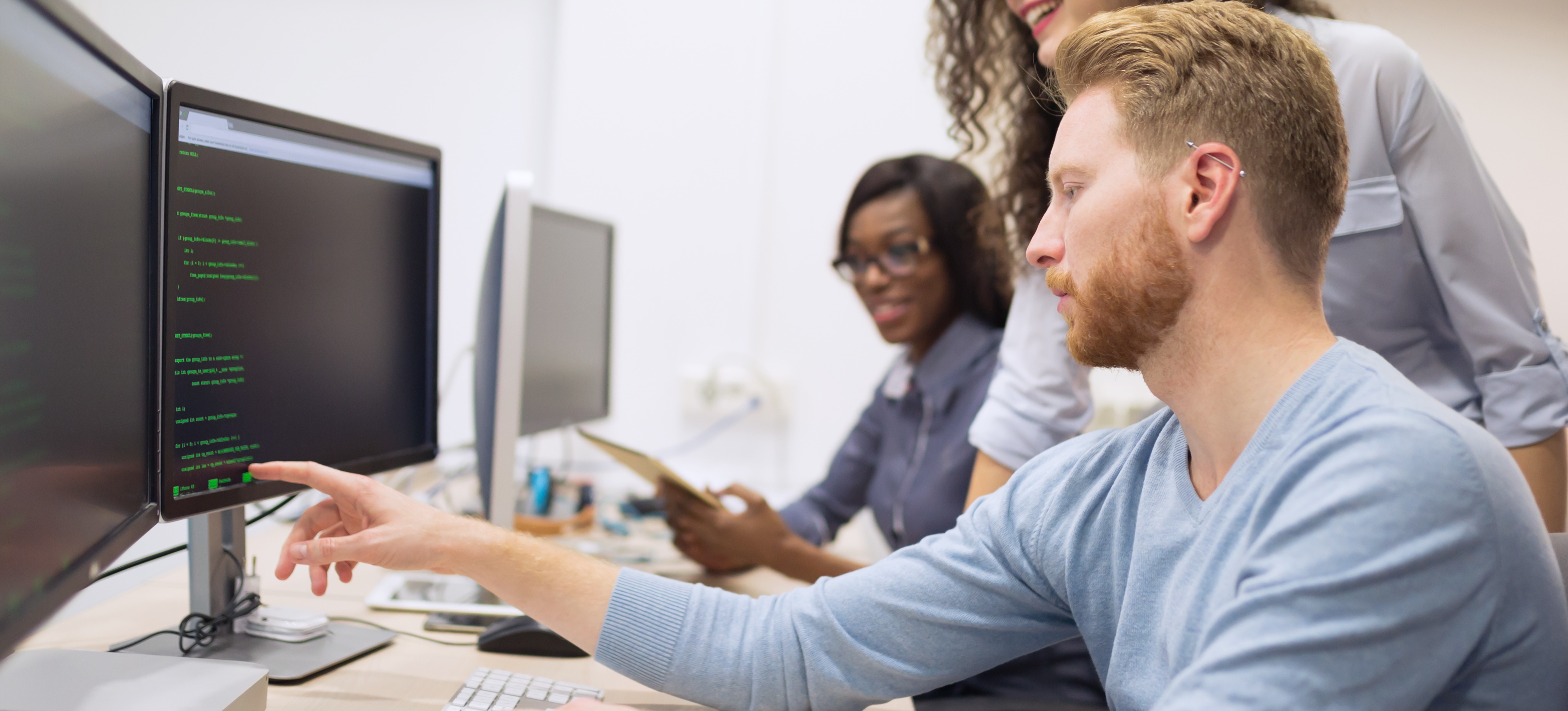 [Featured Image] A seated programmer working on code points to a unique identifier on their desktop computer while talking to two colleagues.
