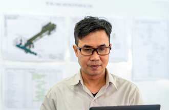 [Featured Image]:  A male, with short brown hair, wearing a white shirt and glasses, is sitting in front of his laptop, sitting behind documents and graphs that are on the wall. 